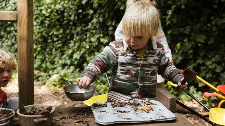 A boy in a knitted jumper is busy playing in the mud pie kitchen, concentrating on what he's doing.